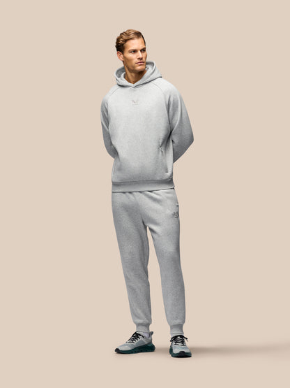 A man modeling a light gray heathered two-piece tracksuit, consisting of a pullover hoodie and matching jogger pants, paired with dark sneakers, against a solid beige background.