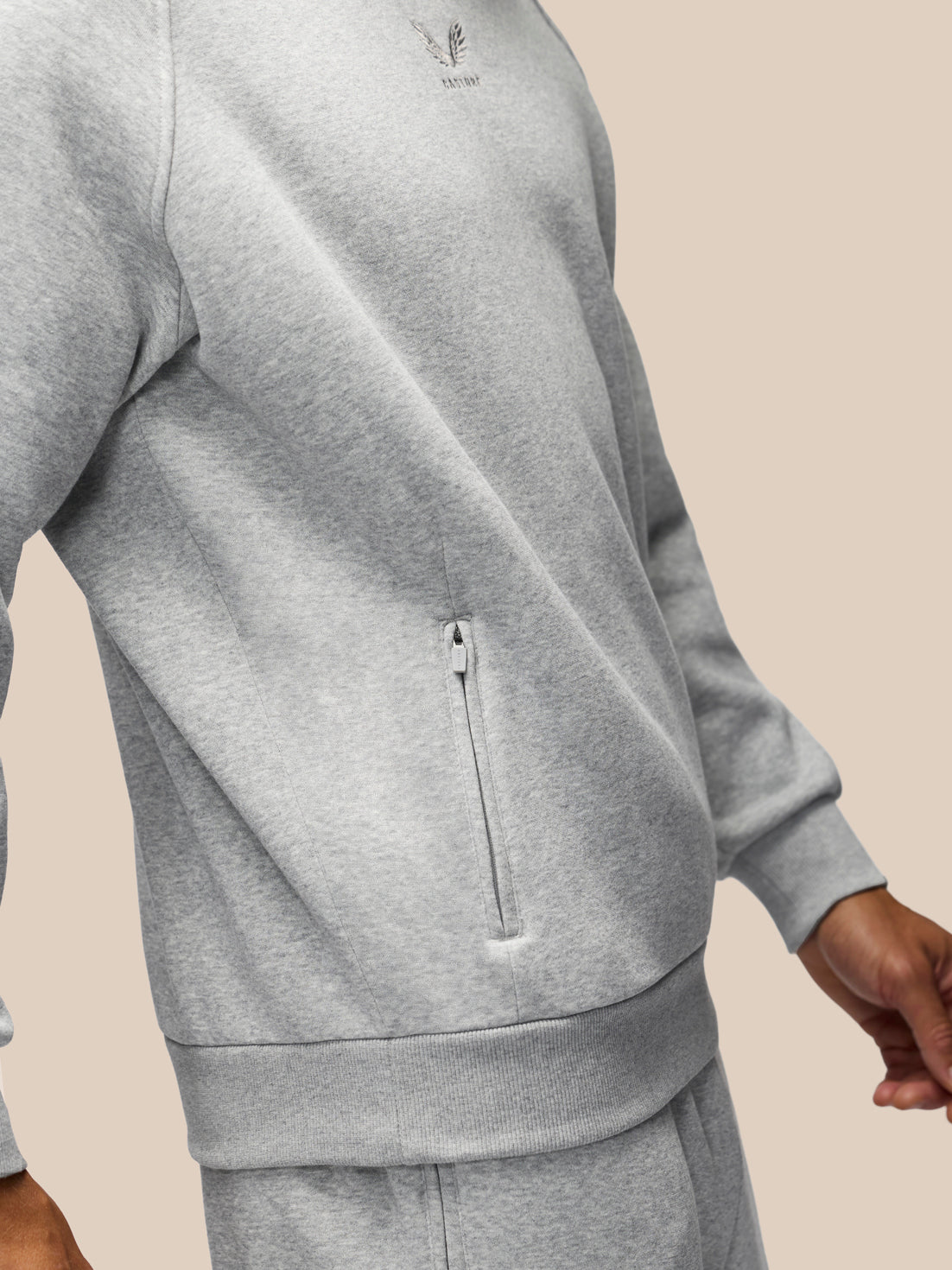 A close-up of a person wearing a light heather gray zippered sweatshirt and matching sweatpants against a solid beige background.