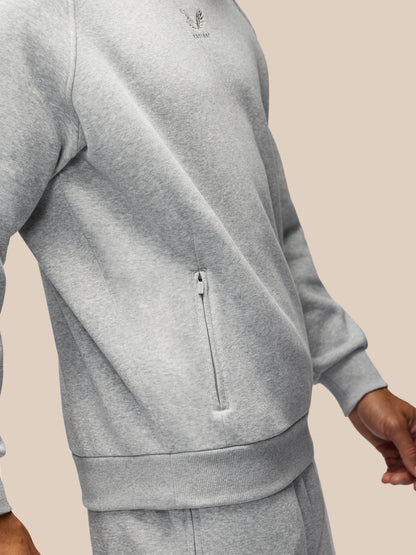 A close-up of a person wearing a light heather gray zippered sweatshirt and matching sweatpants against a solid beige background.
