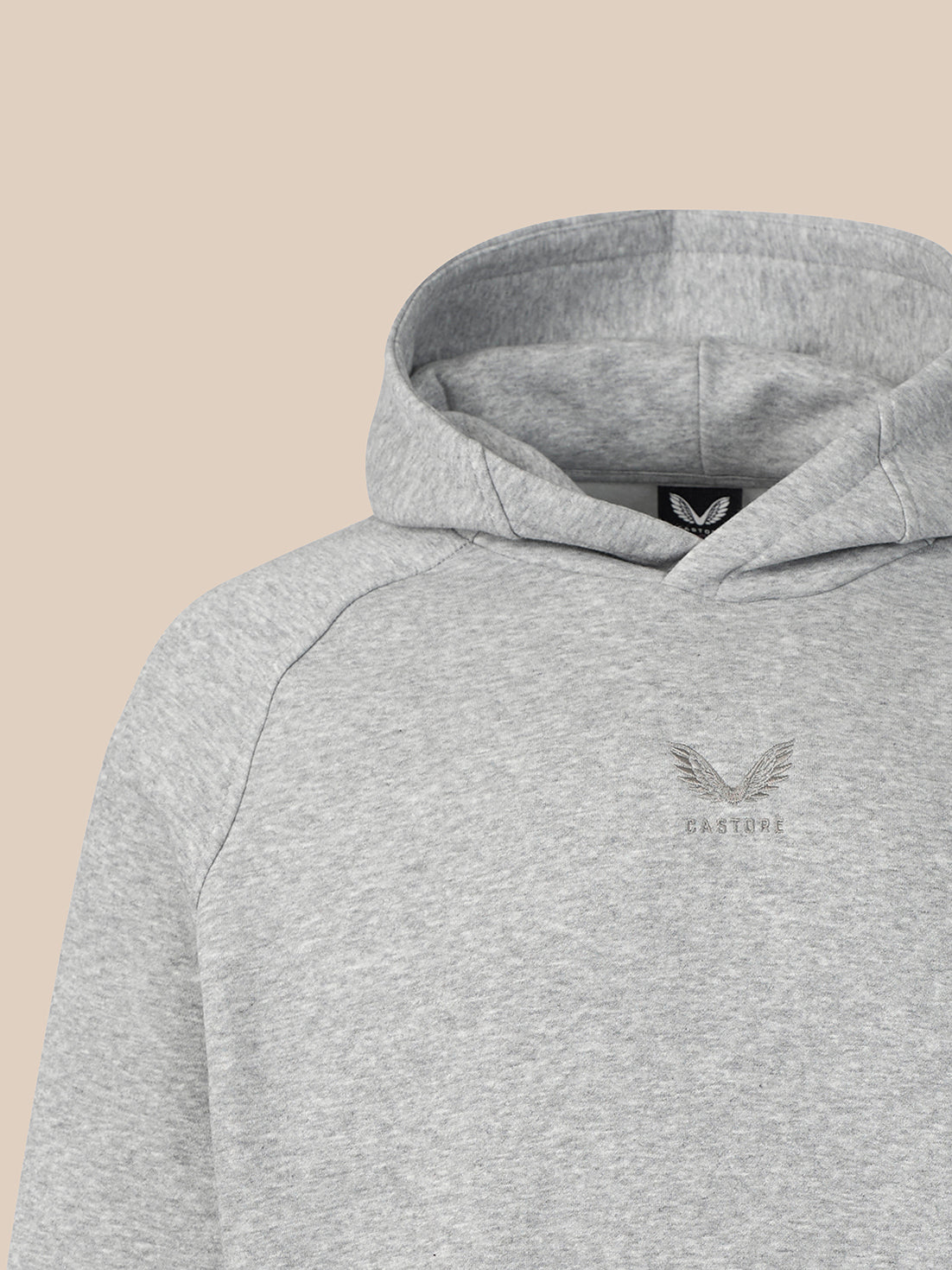 Close-up of a light heather gray pullover hoodie with a small embroidered winged logo and the word 'CASTORE' on the left chest, shown against a solid beige background.