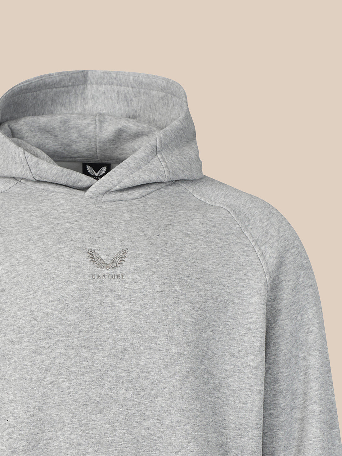 Close-up of a light heather gray pullover hoodie with a small tonal winged logo and the word 'CASTORE' printed on the left chest, set against a solid beige background.