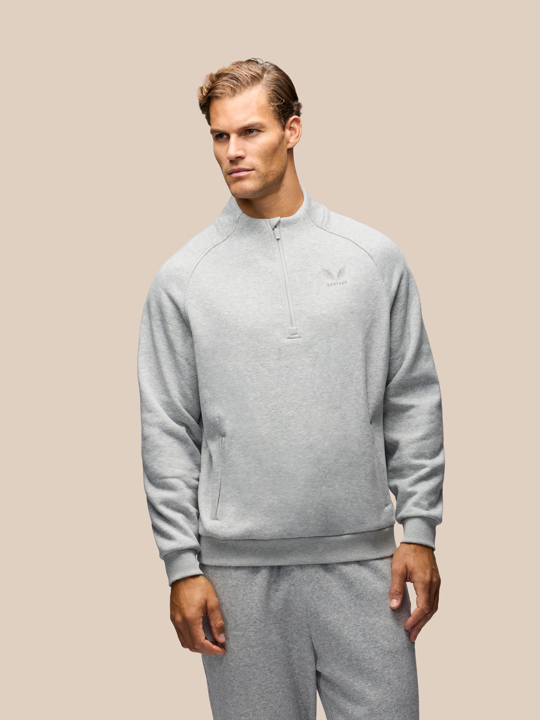 A man wearing a light gray heathered quarter-zip sweatshirt and matching sweatpants, standing against a solid beige background.