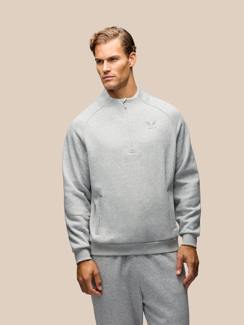 A man wearing a light gray heathered quarter-zip sweatshirt and matching sweatpants, standing against a solid beige background.