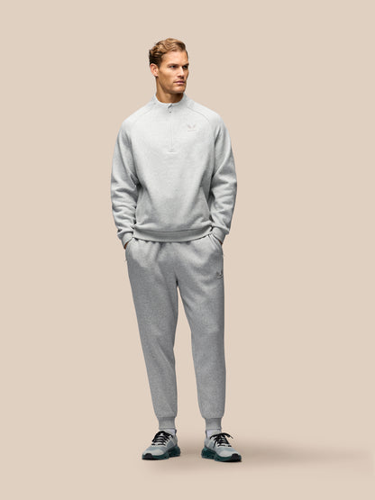 A man modeling a light gray heathered quarter-zip sweatshirt and matching jogger sweatpants against a solid beige background.
