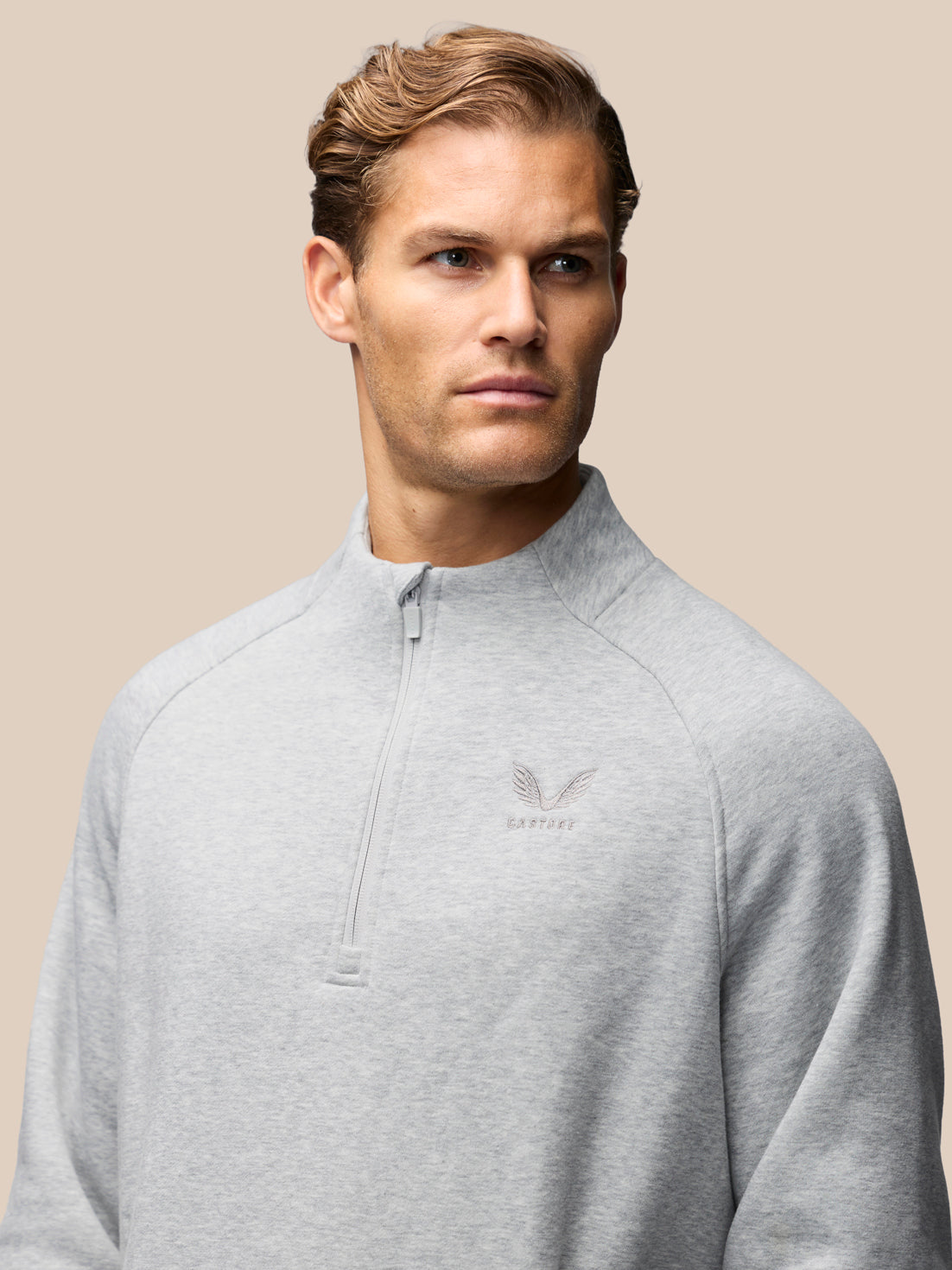 A man wearing a light gray heathered quarter-zip pullover sweatshirt with a small embroidered winged logo on the left chest, set against a solid beige background.