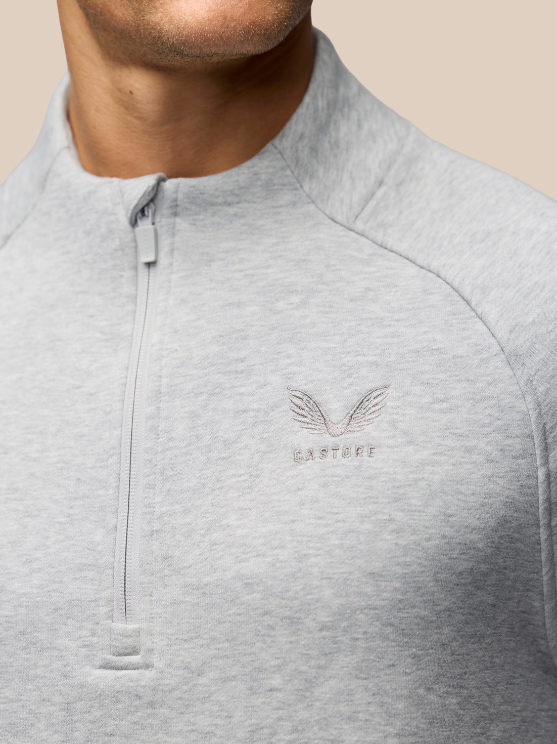 Close-up of a man wearing a light gray heathered quarter-zip pullover sweatshirt with a subtle embroidered winged logo and the word 'CASTORE' on the left chest, against a solid beige background.