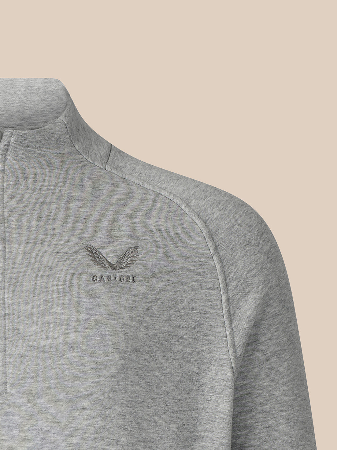 Close-up of a heather gray quarter-zip pullover sweatshirt with a small embroidered winged logo and the word 'CASTORE' on the left chest, set against a solid beige background.