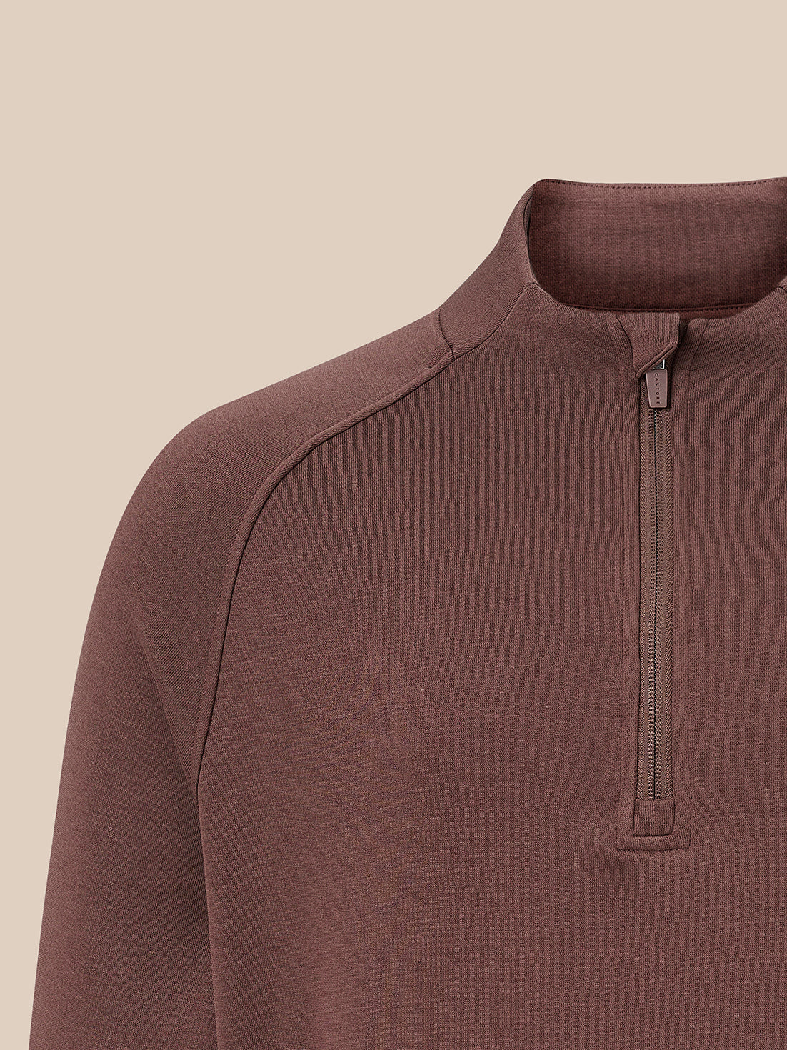 Close-up of a brown quarter-zip pullover sweater on a model, showing the collar, zipper, and shoulder seam against a solid beige background.