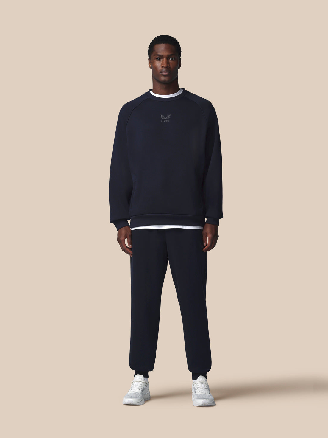 Man wearing a navy blue sweatshirt and pants with white sneakers on a beige background