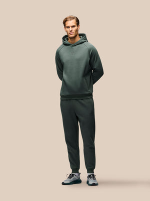 A man modeling a dark green two-piece tracksuit consisting of a pullover hoodie and matching jogger pants, paired with gray and black sneakers, against a solid beige background.
