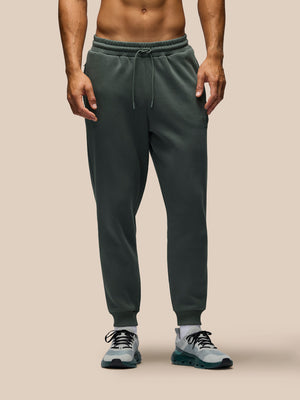 A man wearing dark green jogger sweatpants with a drawstring waist and ribbed cuffs, paired with gray sneakers, shown from the waist down against a solid beige background.