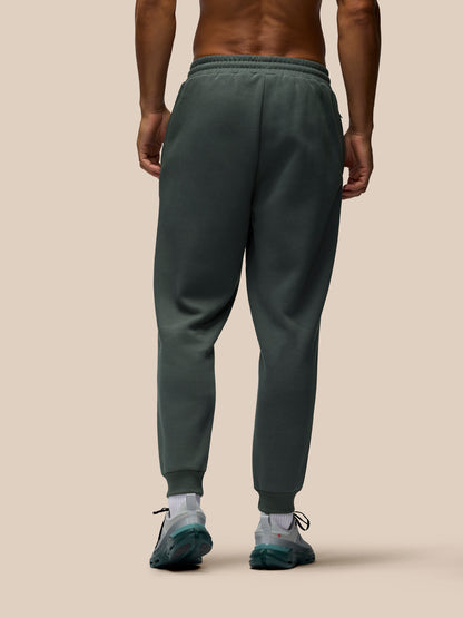 A man viewed from the back wearing dark green sweatpants, white socks, and athletic shoes against a solid beige background.