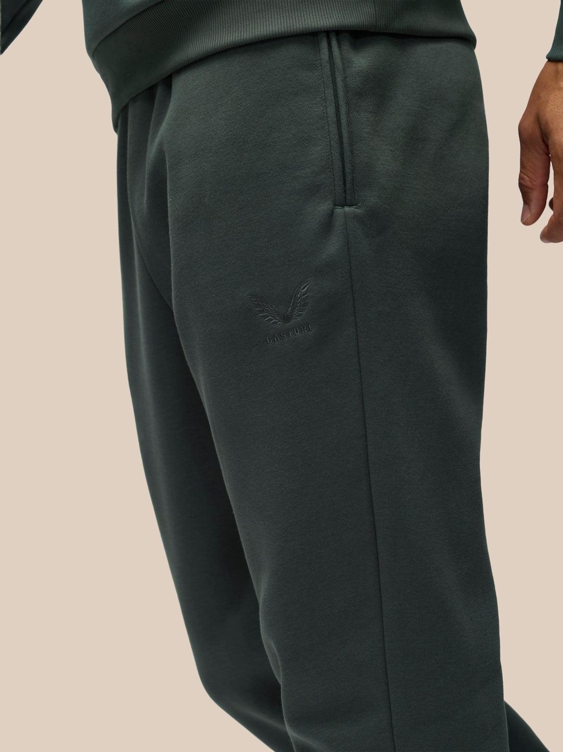 Close-up of a person wearing dark green sweatpants with a tonal embroidered logo on the upper thigh, shown against a plain beige background.