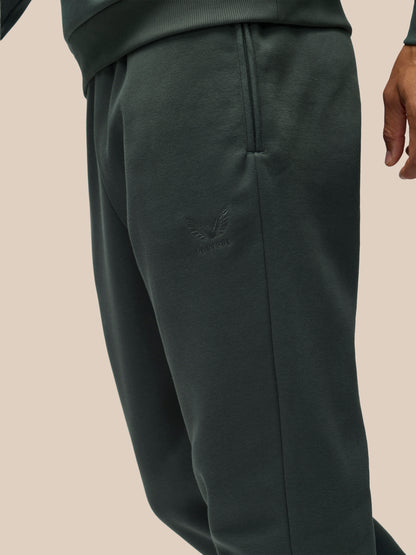 Close-up of a person wearing dark green sweatpants with a tonal embroidered logo on the upper thigh, shown against a plain beige background.