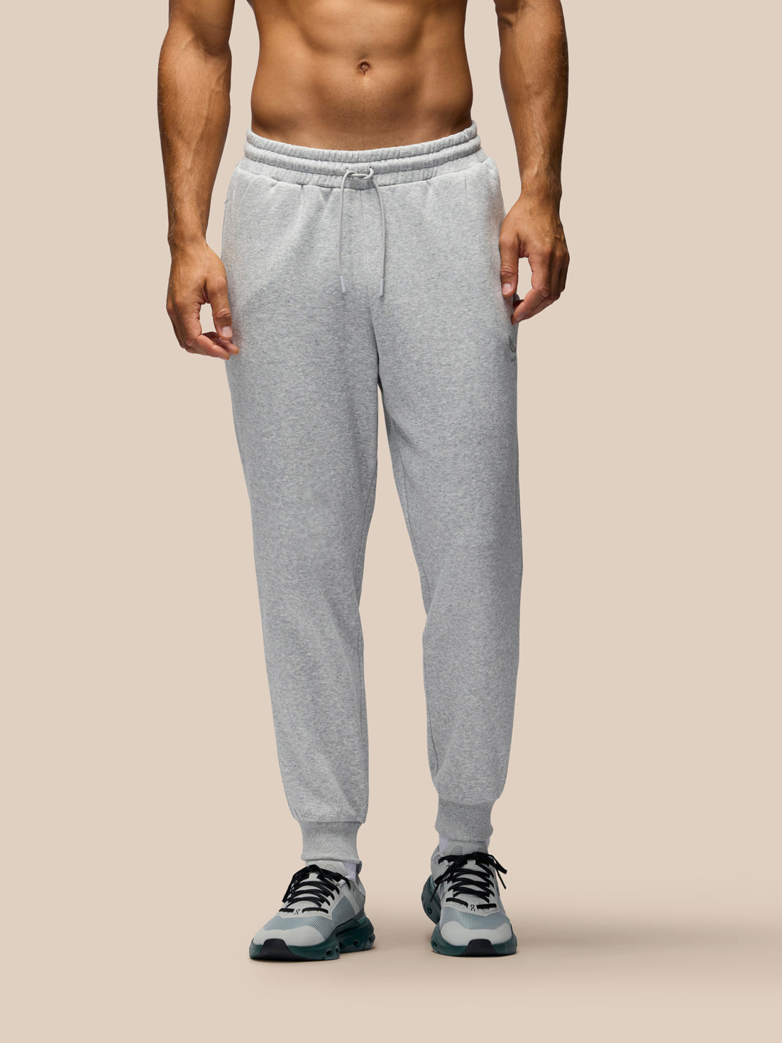 A man wearing light gray heather jogger sweatpants with a drawstring waist and ribbed cuffs, paired with gray athletic sneakers, shown from the waist down against a plain beige studio background.