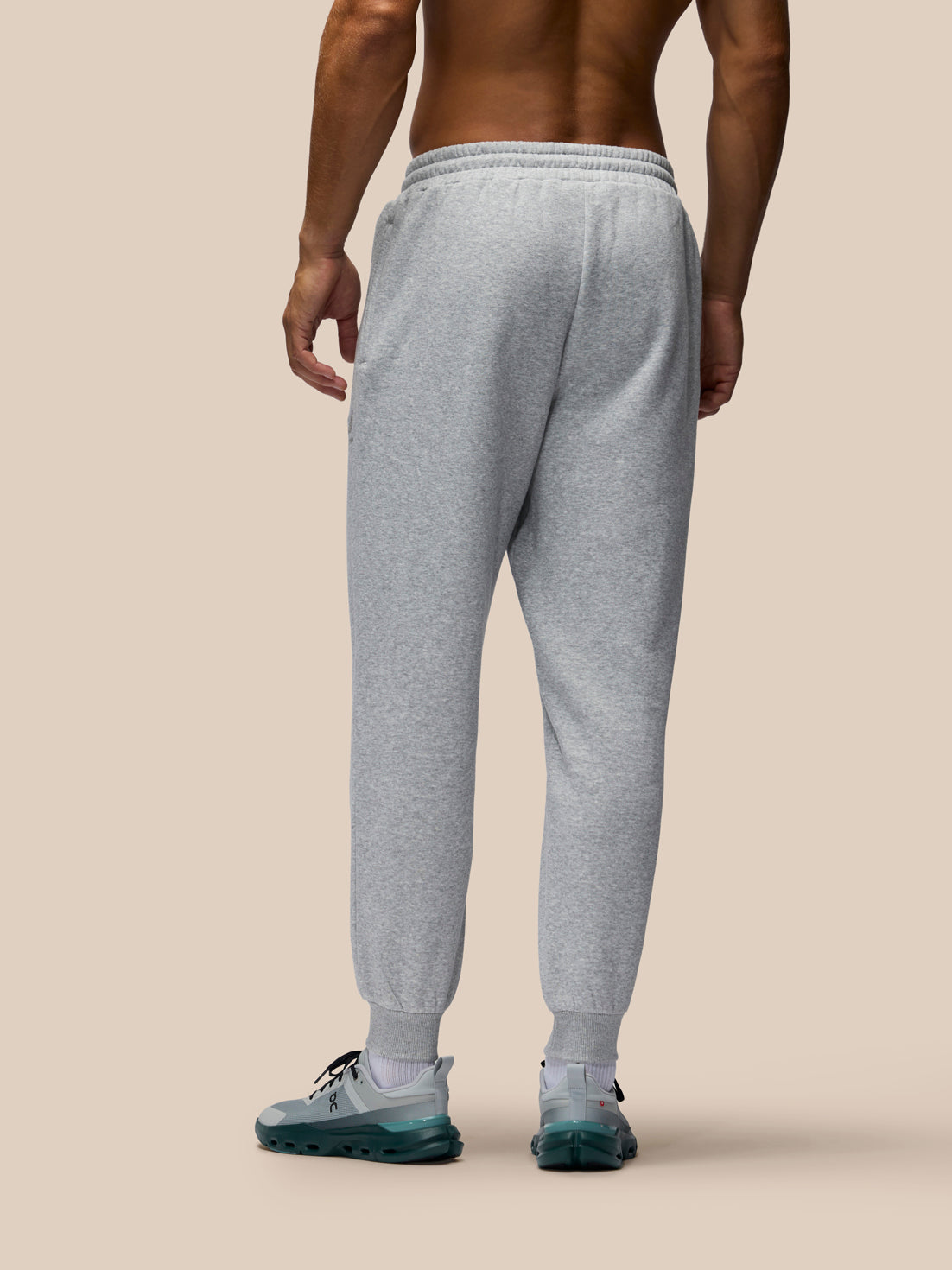 A man shown from the back wearing light heather gray jogger sweatpants and teal sneakers, standing against a solid beige studio background.