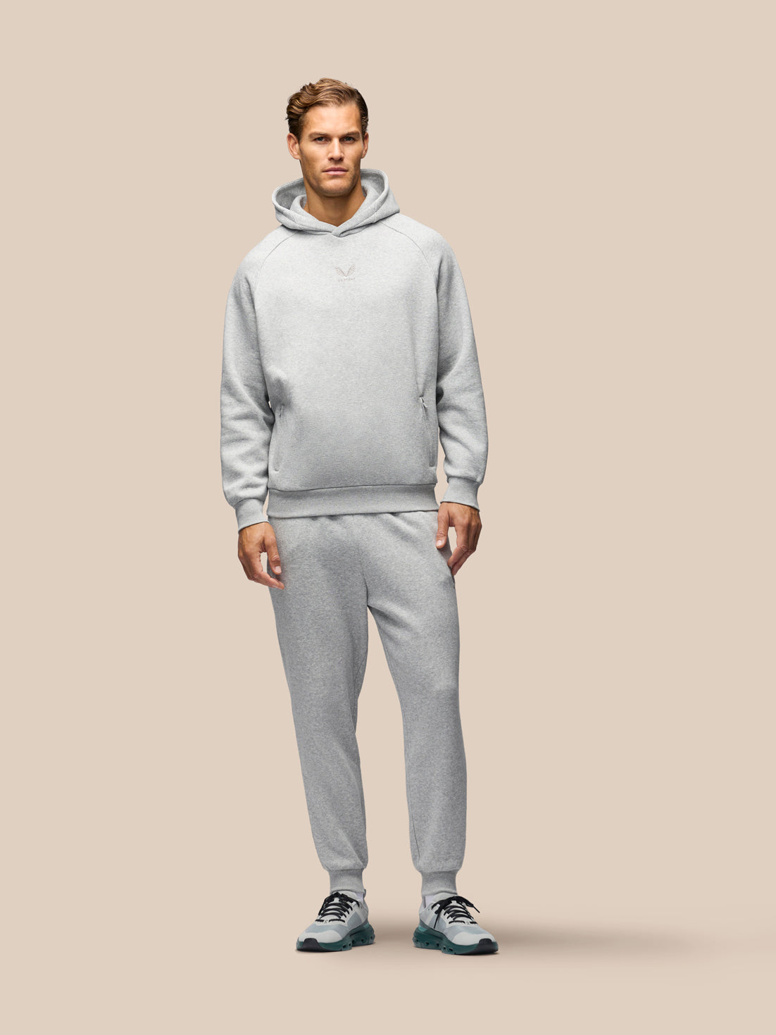 A man modeling a light gray heathered hooded sweatshirt and matching jogger sweatpants set against a solid beige background.