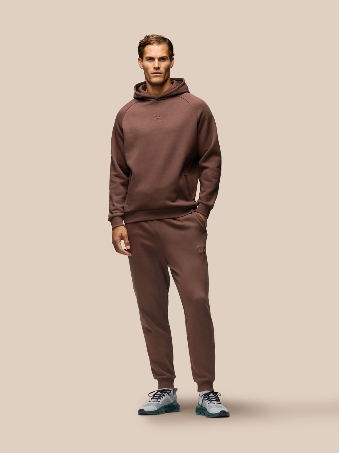 A man modeling a matching brown hoodie and sweatpants set, standing against a solid beige background.
