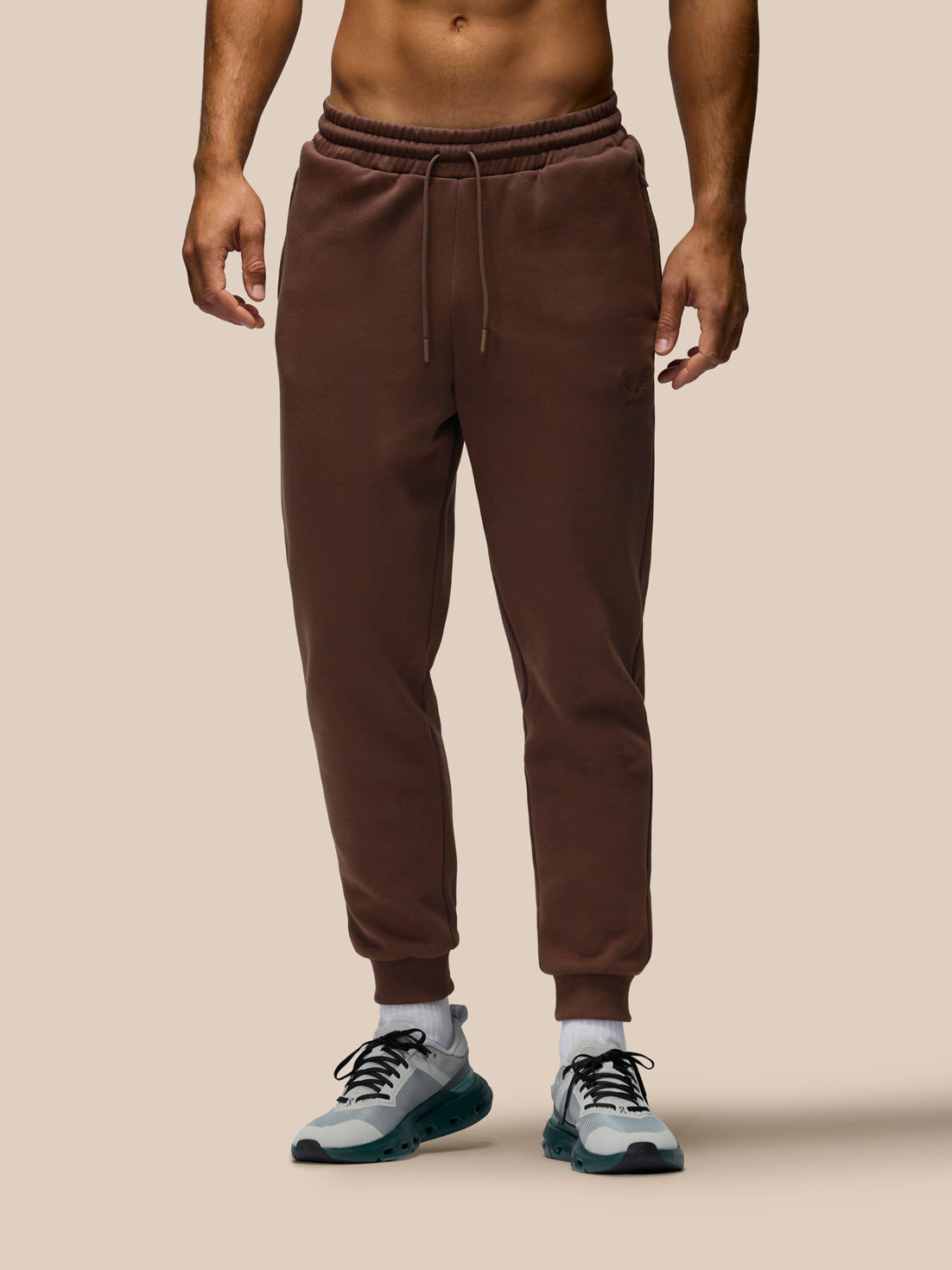 A man wearing brown jogger sweatpants with a drawstring waist and elastic cuffs, paired with gray sneakers, shown from the waist down against a solid beige background.