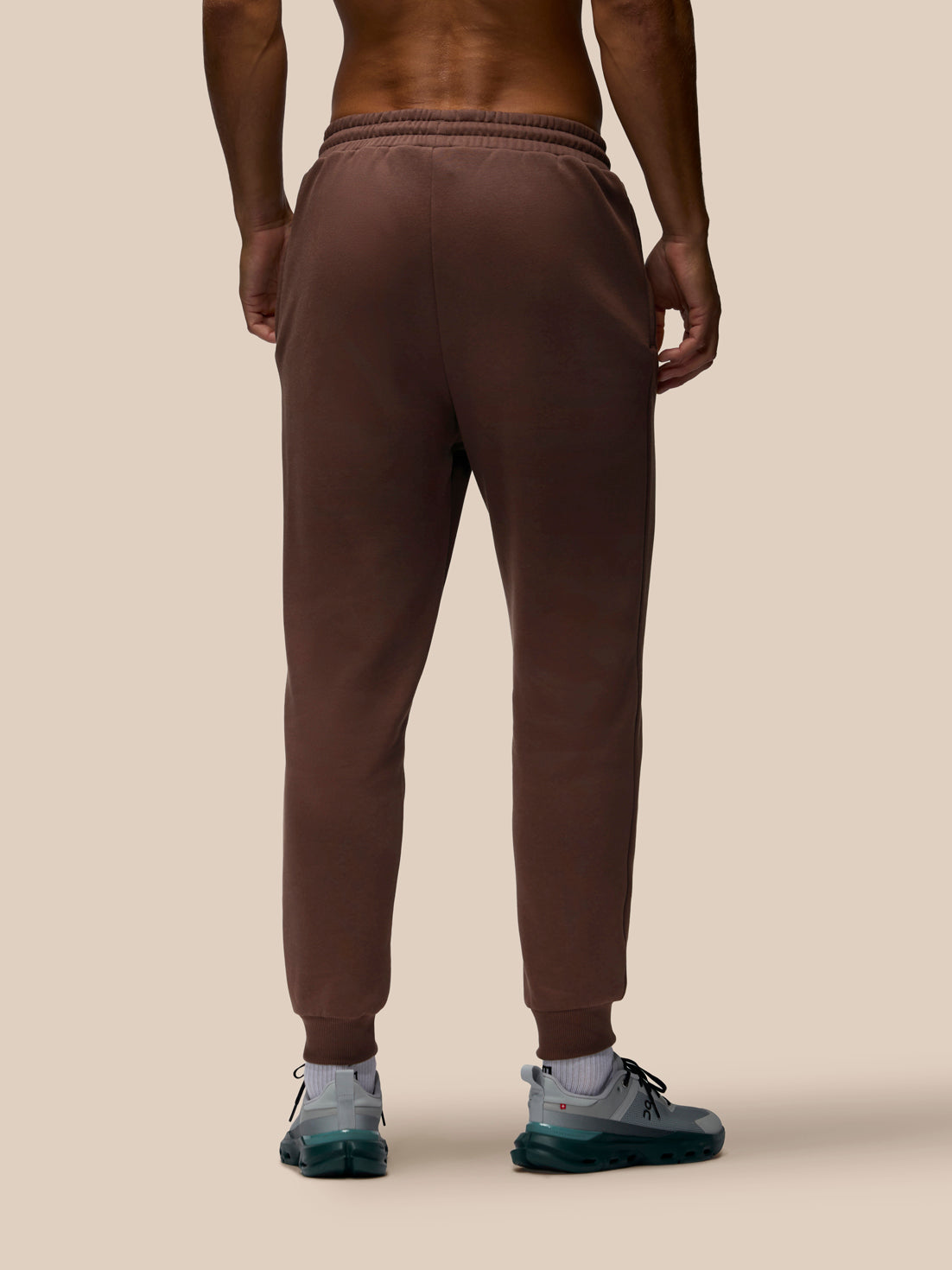 Rear view of a person wearing brown jogger sweatpants with elastic waistband and ribbed cuffs, paired with white socks and gray sneakers, against a plain beige studio background.