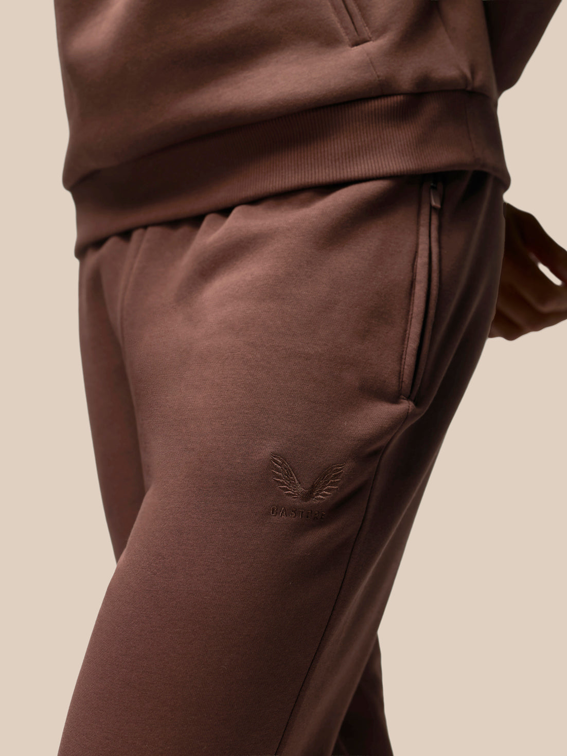 Close-up of a person wearing brown sweatpants with a subtle tonal embroidered logo on the thigh, paired with a matching brown top against a plain beige background.