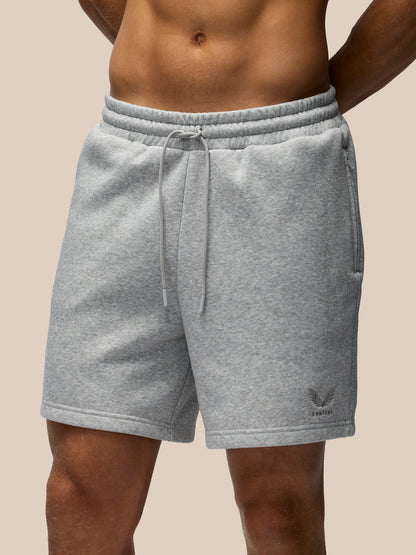 A close-up of a person wearing light heather gray drawstring shorts against a plain beige background. The shorts have an elastic waistband, side pockets, and a small dark logo near the lower left leg.