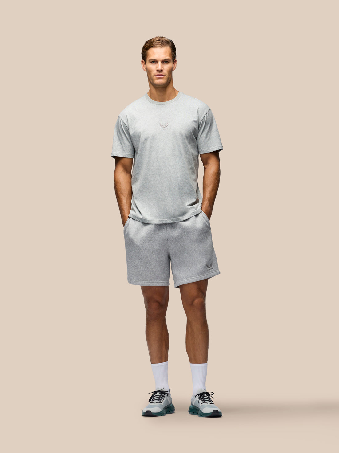 A man modeling a light gray heathered athletic t-shirt and matching shorts against a solid beige background.