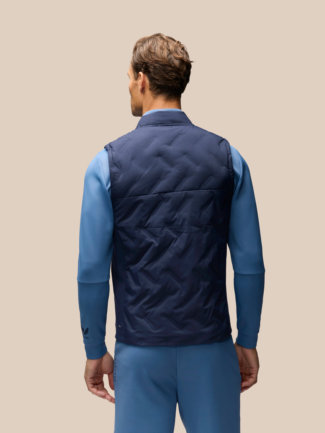A man shown from the back wearing a navy blue quilted puffer vest over a blue long-sleeve shirt and matching blue pants, against a solid beige background.