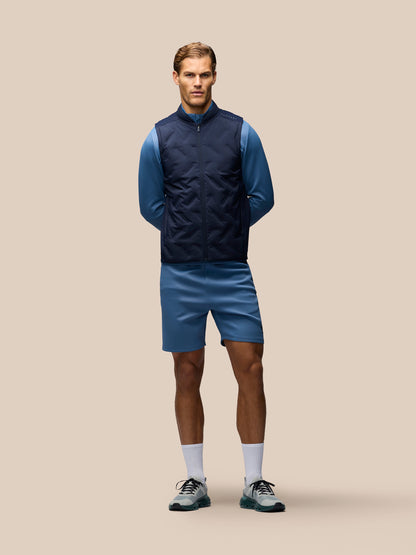 A male model stands against a solid beige studio background wearing a navy blue zip-up puffer vest over a blue long-sleeve shirt, blue athletic shorts, white socks, and dark athletic shoes.