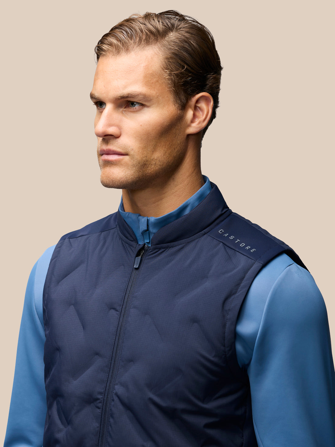 A man wearing a navy blue quilted zip-up vest over a blue long-sleeve shirt, shown in a close-up profile shot against a solid beige background. The brand name 'CASTORE' is visible on the shoulder.