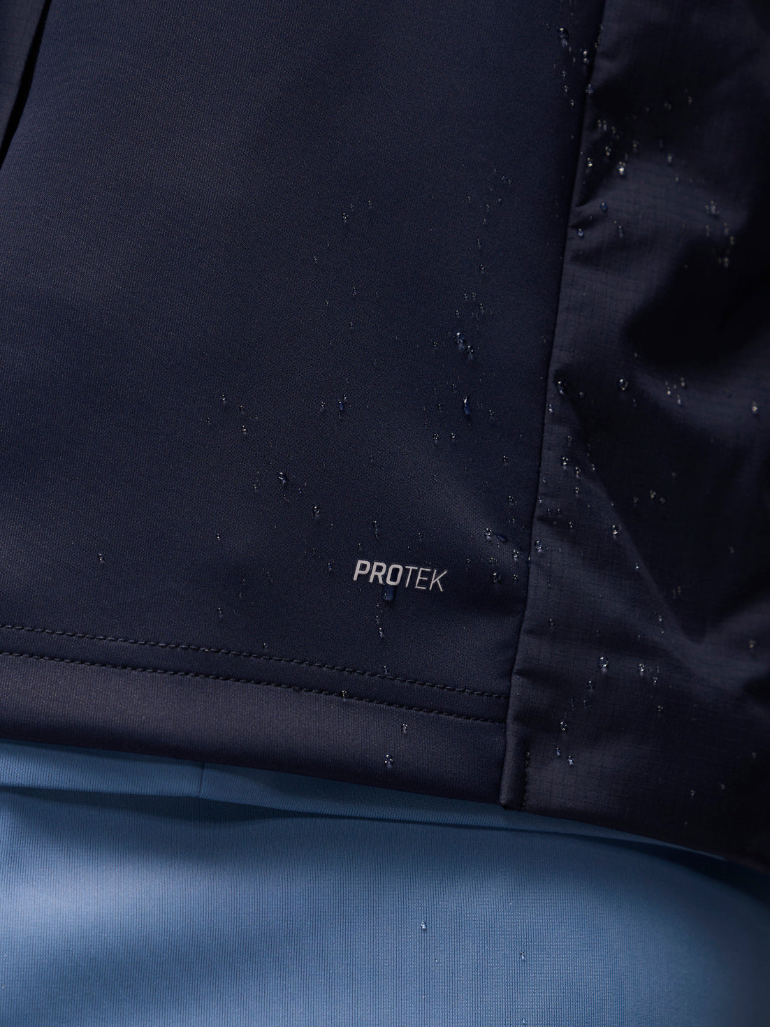 Close-up of a dark navy waterproof jacket fabric with water droplets on the surface and a small 'PROTEK' logo near the hem, set against a blue background.