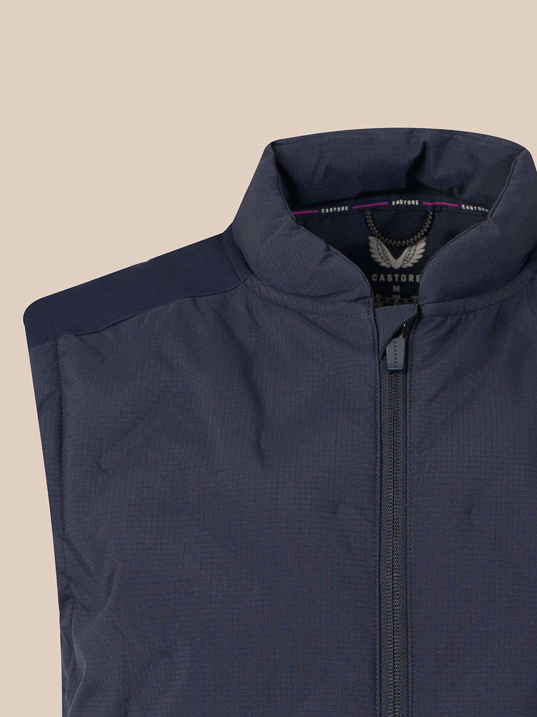 Close-up of a navy blue zip-up vest with a stand-up collar and shoulder yoke, shown against a solid beige background. The brand label inside the collar reads CASTORE.