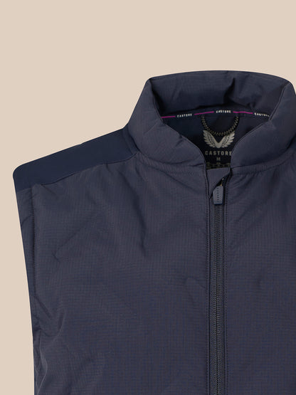 Close-up of a navy blue zip-up vest with a stand-up collar and shoulder yoke, shown against a solid beige background. The brand label inside the collar reads CASTORE.