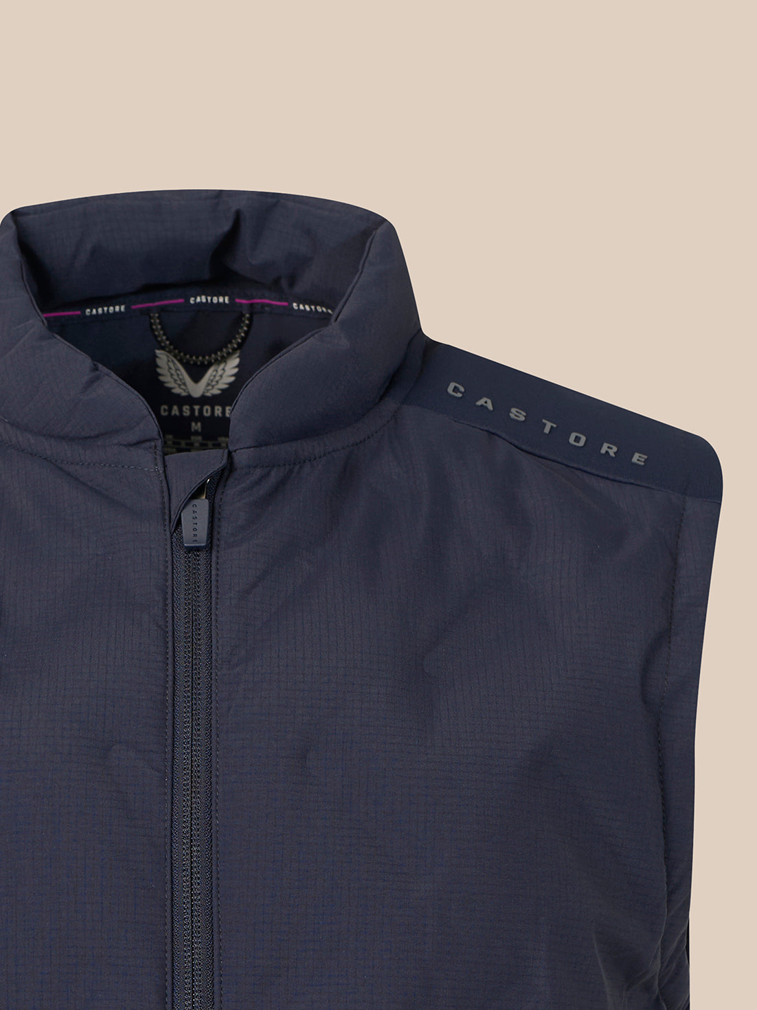 Close-up of a Castore lightweight zip-up vest in dark navy blue, showing the collar, front zipper, and the Castore brand name printed on the shoulder against a solid beige background.