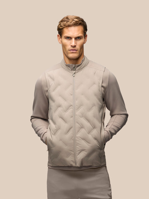 A man wearing a light taupe-beige zip-up quilted vest over a matching long-sleeve top and pants, photographed against a solid light beige background.