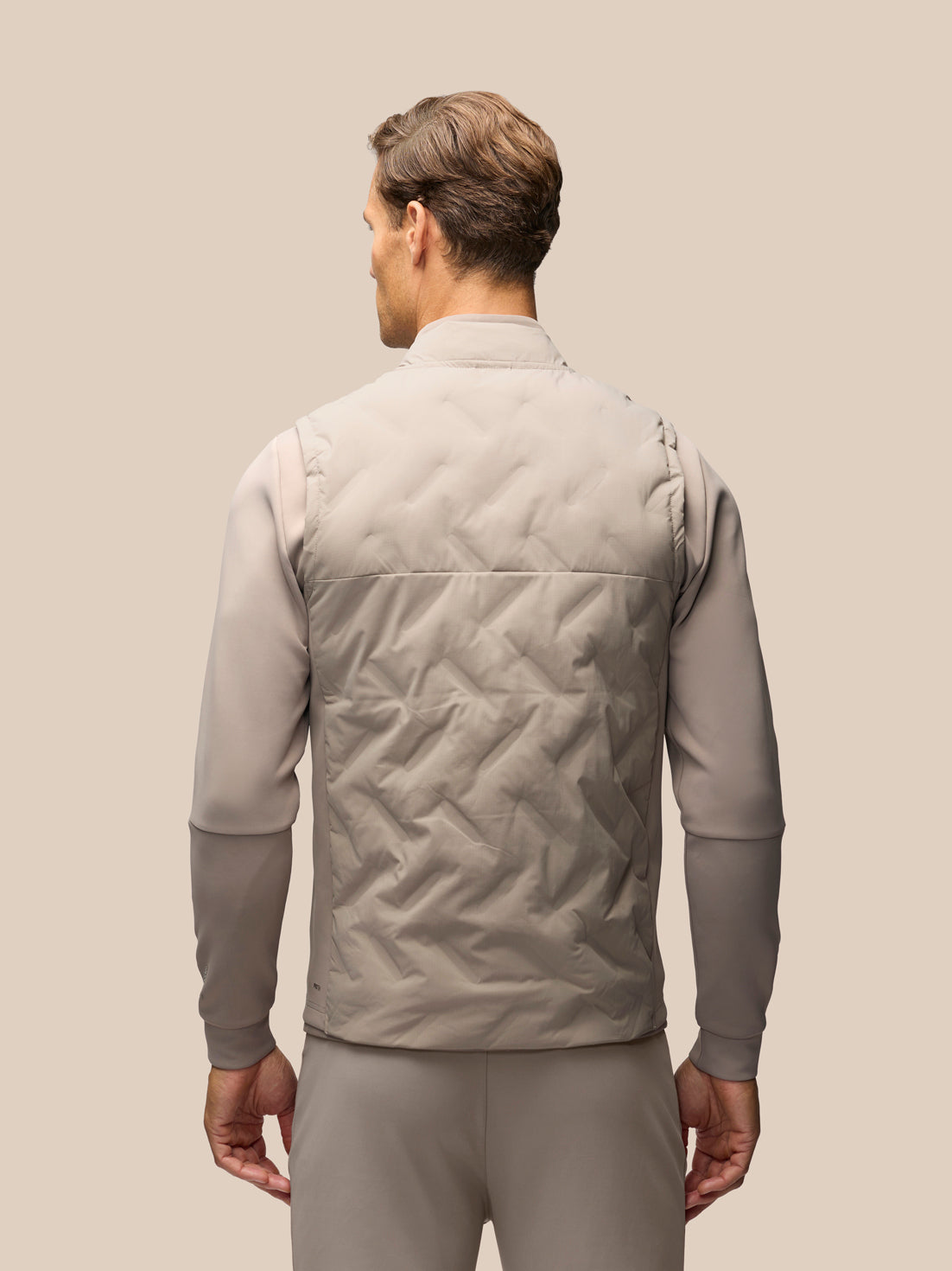 A man shown from the back wearing a light taupe quilted vest over a matching long-sleeve shirt and trousers, set against a solid beige background.