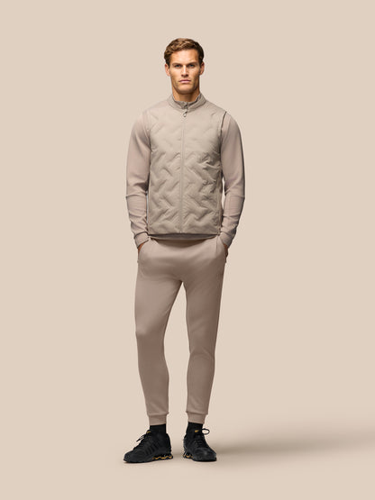 A male model stands against a solid beige studio background wearing a matching beige tracksuit consisting of a zip-up quilted jacket, a long-sleeve knit top, and slim-fit pants, paired with black athletic shoes.