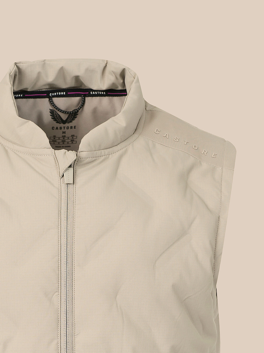 Close-up of a sleeveless quilted vest in a light warm gray-beige color, featuring a high stand-up collar, front zipper, and subtle 'CASTORE' branding on the shoulder.