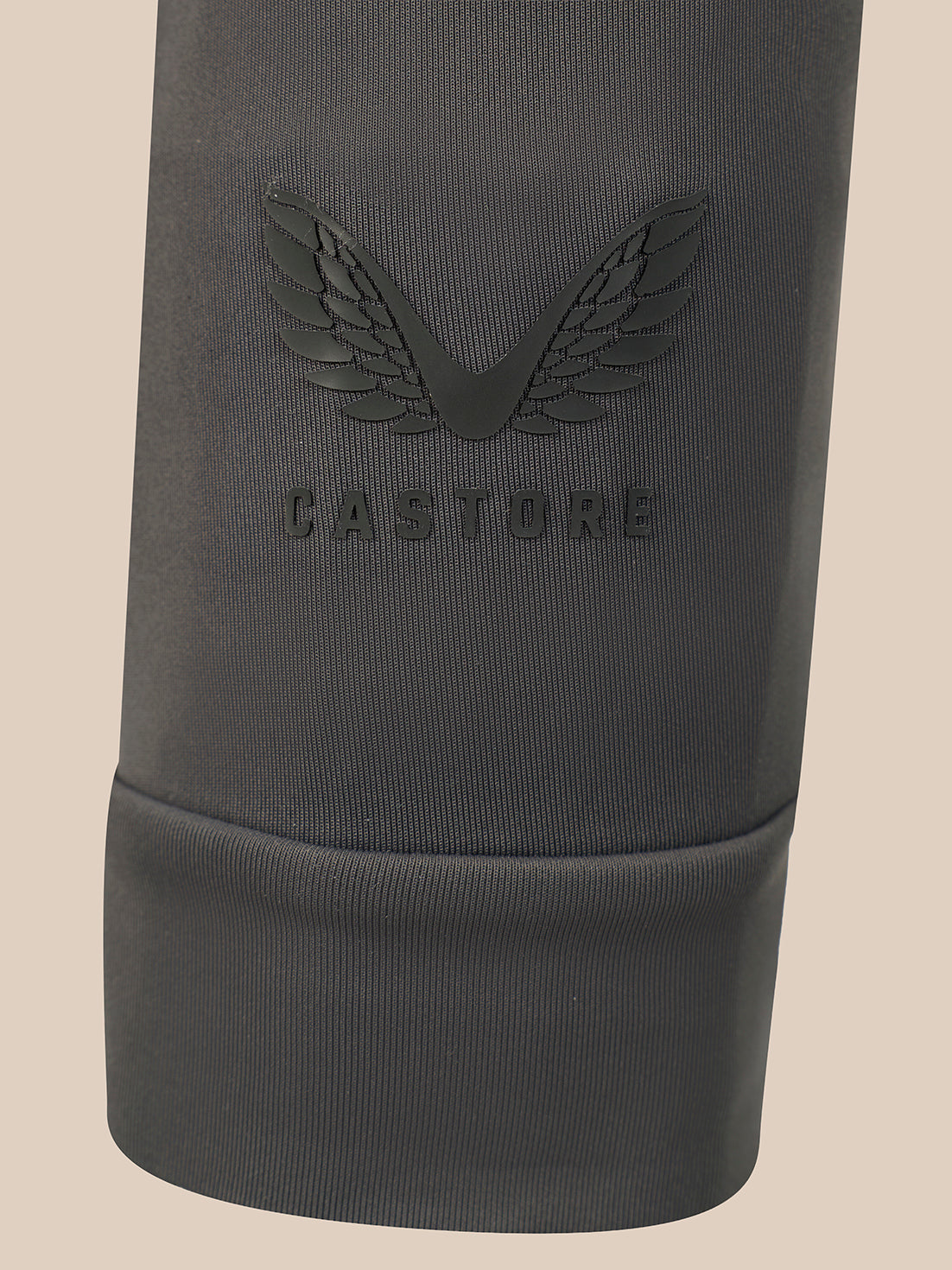 Close-up of a dark gray ribbed fabric sleeve or cuff with an embossed winged logo and the word CASTORE visible on the front, against a plain light beige background.