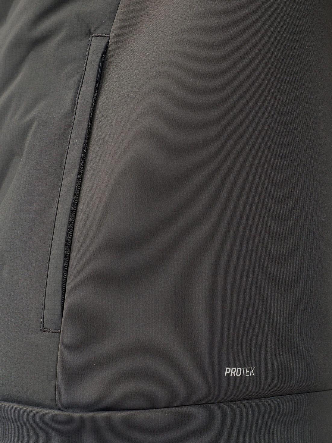 Close-up of a dark gray technical jacket fabric with a vertical zip pocket and a small white PROTEK logo near the bottom.