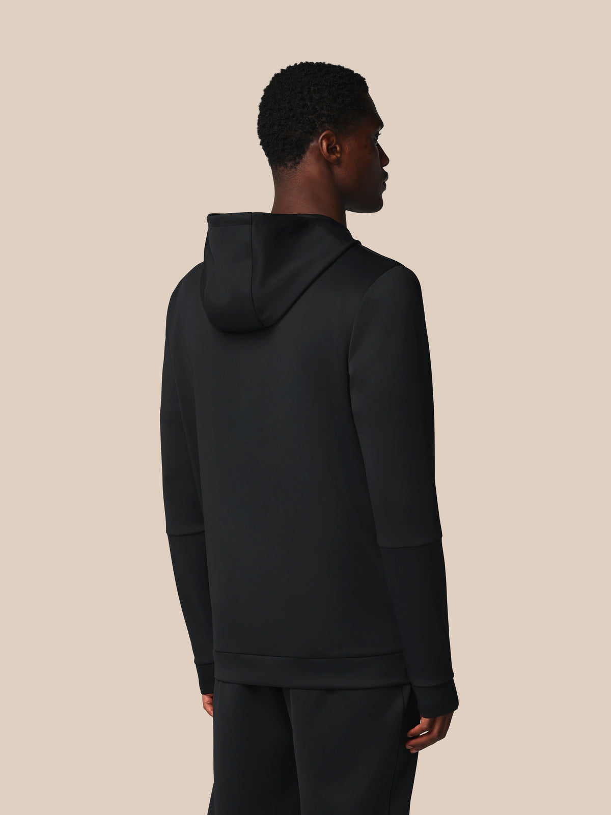 A man shown from the back wearing a black pullover hoodie and matching black pants against a solid beige background.