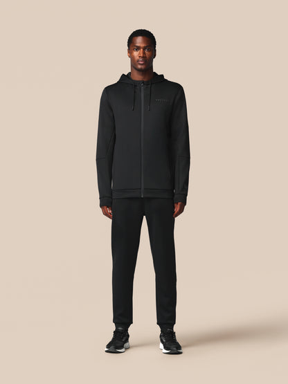 A man modeling a black zip-up hoodie and matching black sweatpants against a solid beige studio background.