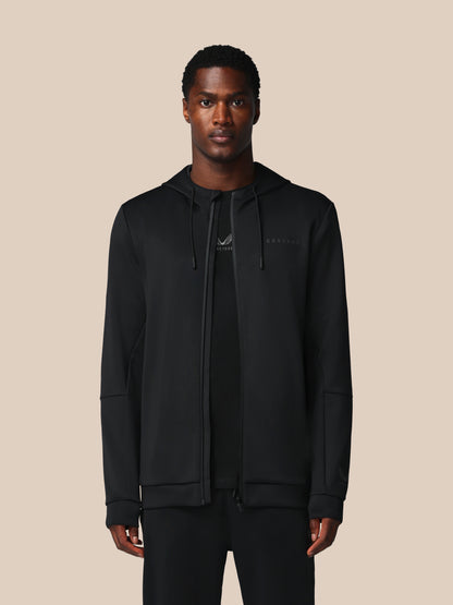 A man modeling a black full-zip hoodie with a drawstring hood and a subtle tonal logo on the left chest, paired with a black t-shirt and black pants, set against a solid beige background.