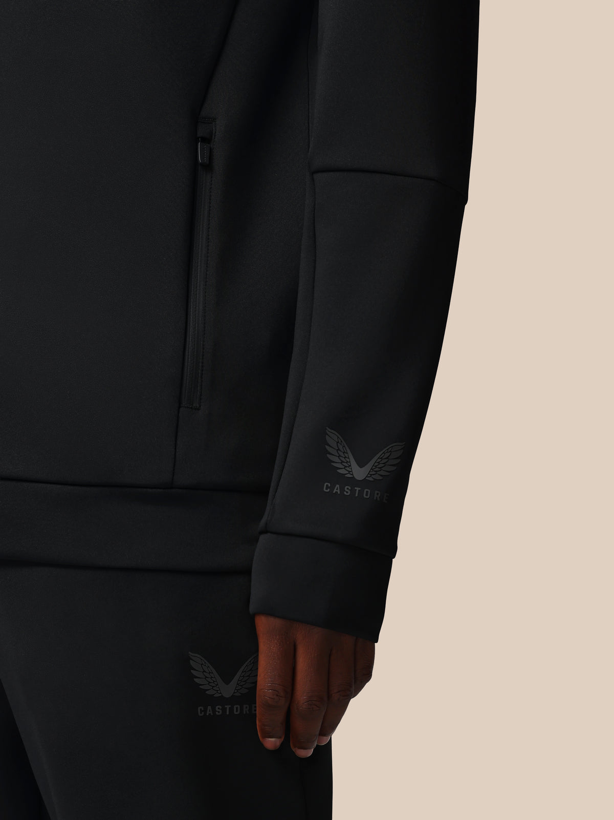 Close-up of a person wearing a dark charcoal zip-front jacket or hoodie with a subtle embroidered winged logo and the word 'CASTORE' on the left sleeve, against a plain warm beige background.