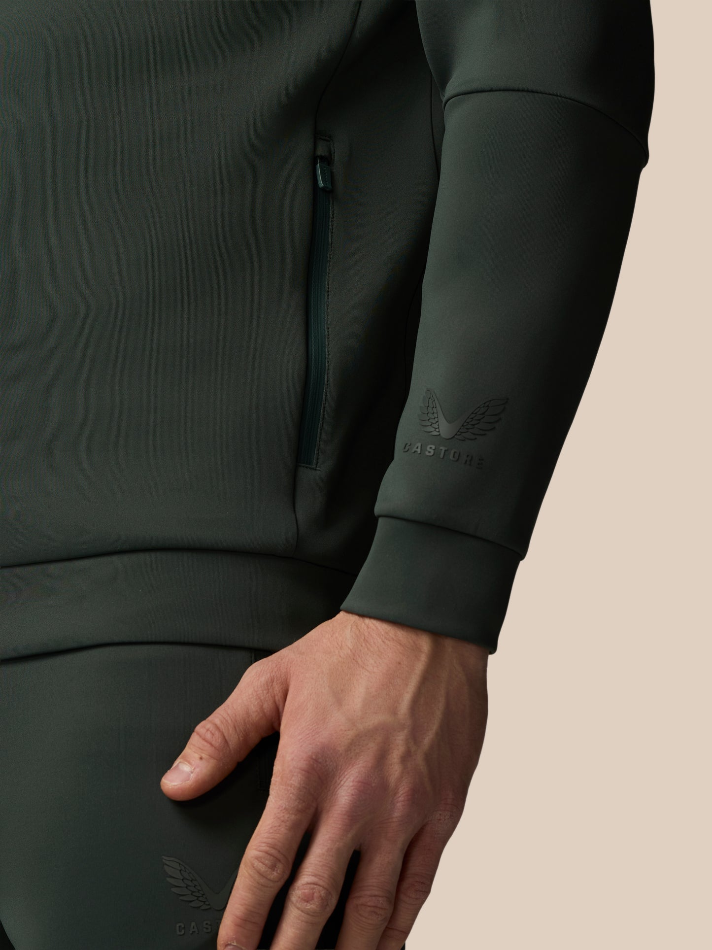Close-up of a person wearing a dark green CASTORE tracksuit with a beige background