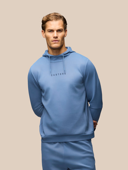 A man wearing a light blue hooded sweatshirt and matching sweatpants against a solid beige background. The hoodie features the brand name 'CASTORE' printed in black on the chest.