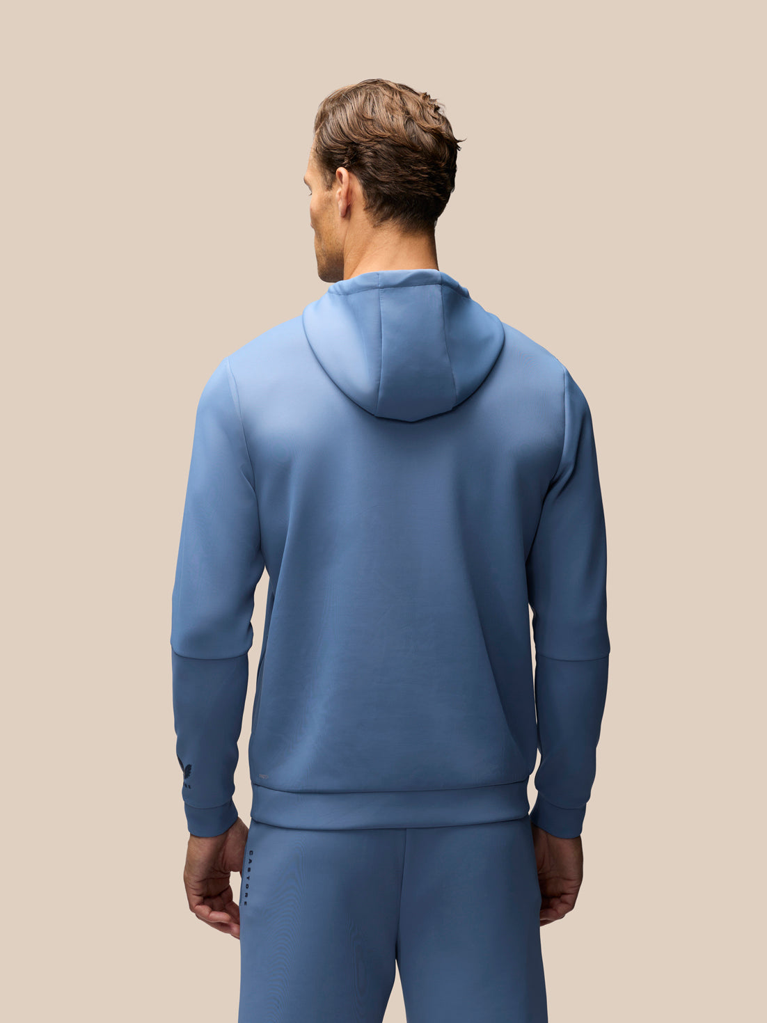 A man viewed from behind wearing a matching dusty blue hoodie and sweatpants set against a solid beige background.
