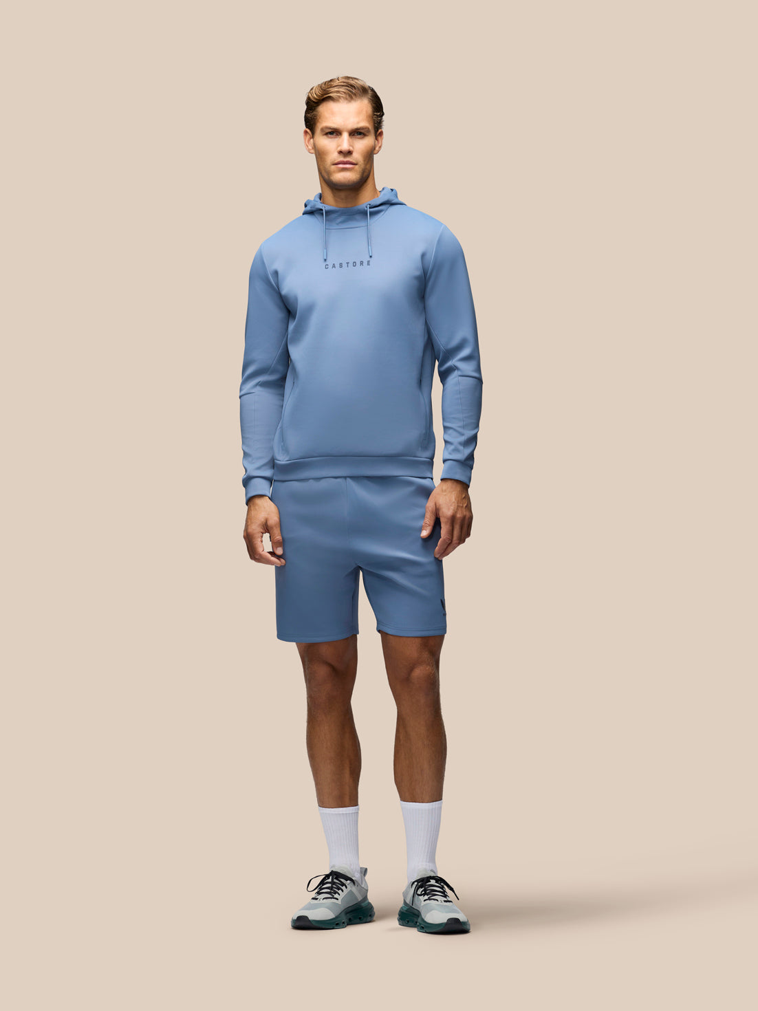 A male model stands against a solid beige background wearing a matching light blue athletic hoodie and shorts set, white socks, and gray running shoes.