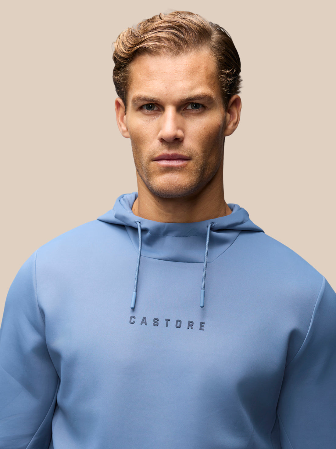 A man wearing a light blue Castore pullover hoodie with 'CASTORE' printed in dark gray letters on the chest, set against a solid beige background.