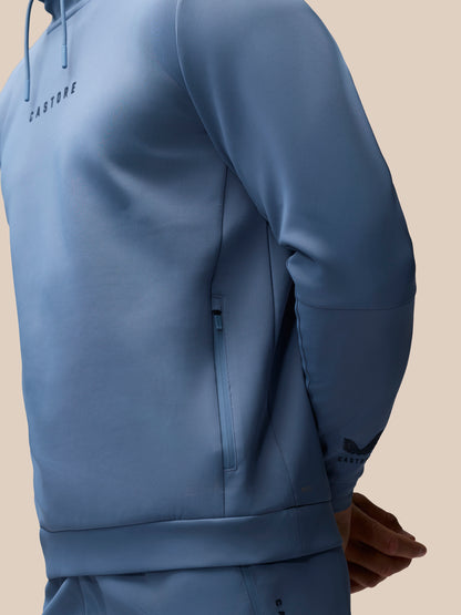 A close-up of a person wearing a slate blue athletic hoodie and matching pants against a solid beige background. The hoodie features a subtle tonal logo on the left chest and a zippered pocket on the side.